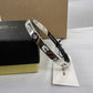 A silver-tone Coach bangle bracelet with three signature lock charms, displayed on a white surface next to its black and gold presentation box and a white brand tag.