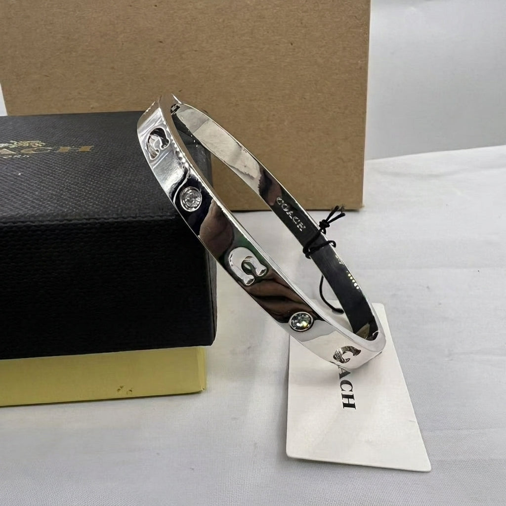 A silver-tone Coach bangle bracelet with three signature lock charms, displayed on a white surface next to its black and gold presentation box and a white brand tag.