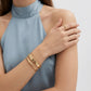 A close-up of a model wearing gold jewelry, featuring a gold bangle bracelet with a central 'G' charm and a matching gold ring, paired with a light blue satin halter top.