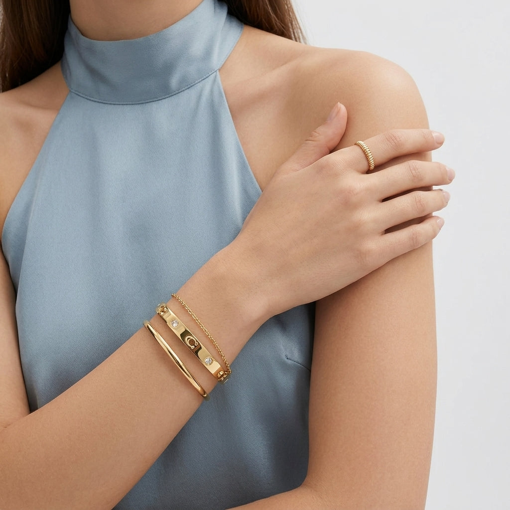 A close-up of a model wearing gold jewelry, featuring a gold bangle bracelet with a central 'G' charm and a matching gold ring, paired with a light blue satin halter top.