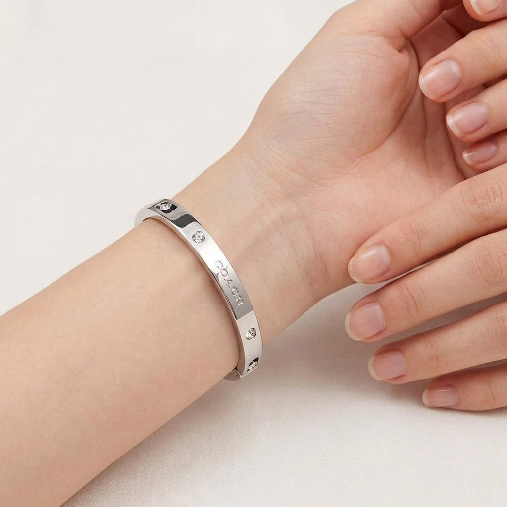 A polished silver-toned metal bangle bracelet with engraved details and small clear stones, shown worn on a person's wrist with their hand resting nearby against a clean white background.