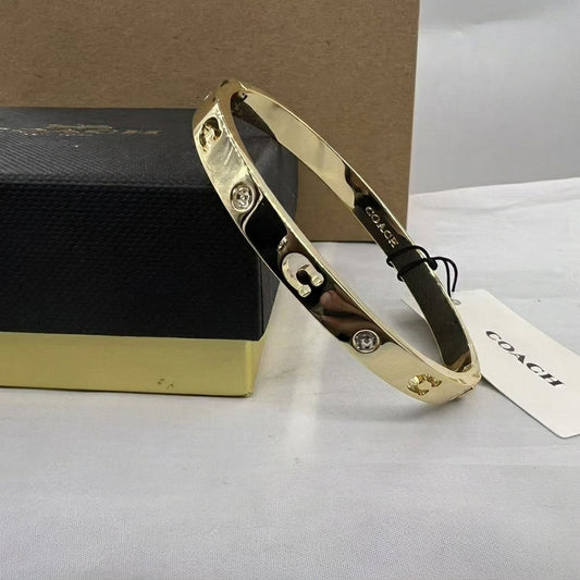 A gold-tone Coach bangle bracelet with a black inlay and signature lock logo, displayed next to its black and gold presentation box and a white COACH tag on a light gray surface.