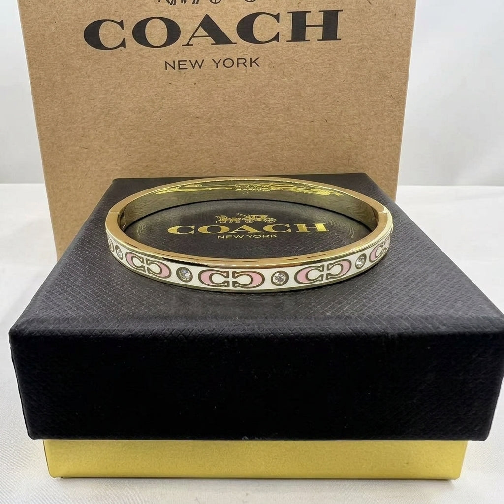 A Coach New York bangle bracelet displayed on a black presentation box with its branded brown box behind it. The bracelet has a gold-tone band with pink and white Coach logo detailing.