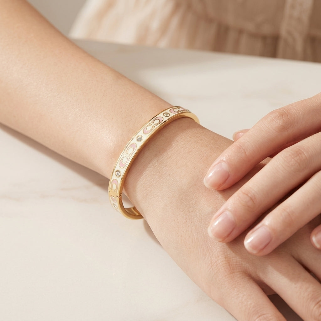 A gold bangle bracelet with engraved floral patterns and small clear stones, shown worn on a person's wrist with their other hand resting on their knee.