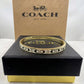 A gold-tone Coach bangle bracelet with signature C logo and small clear stones, displayed on a black Coach box with a brown Coach New York box behind it.