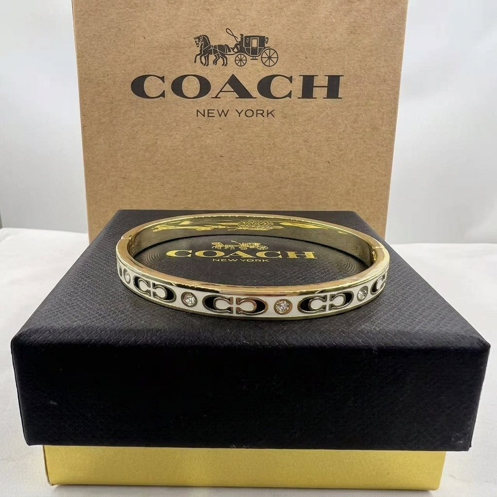 A gold-tone Coach bangle bracelet with signature C logo and small clear stones, displayed on a black Coach box with a brown Coach New York box behind it.