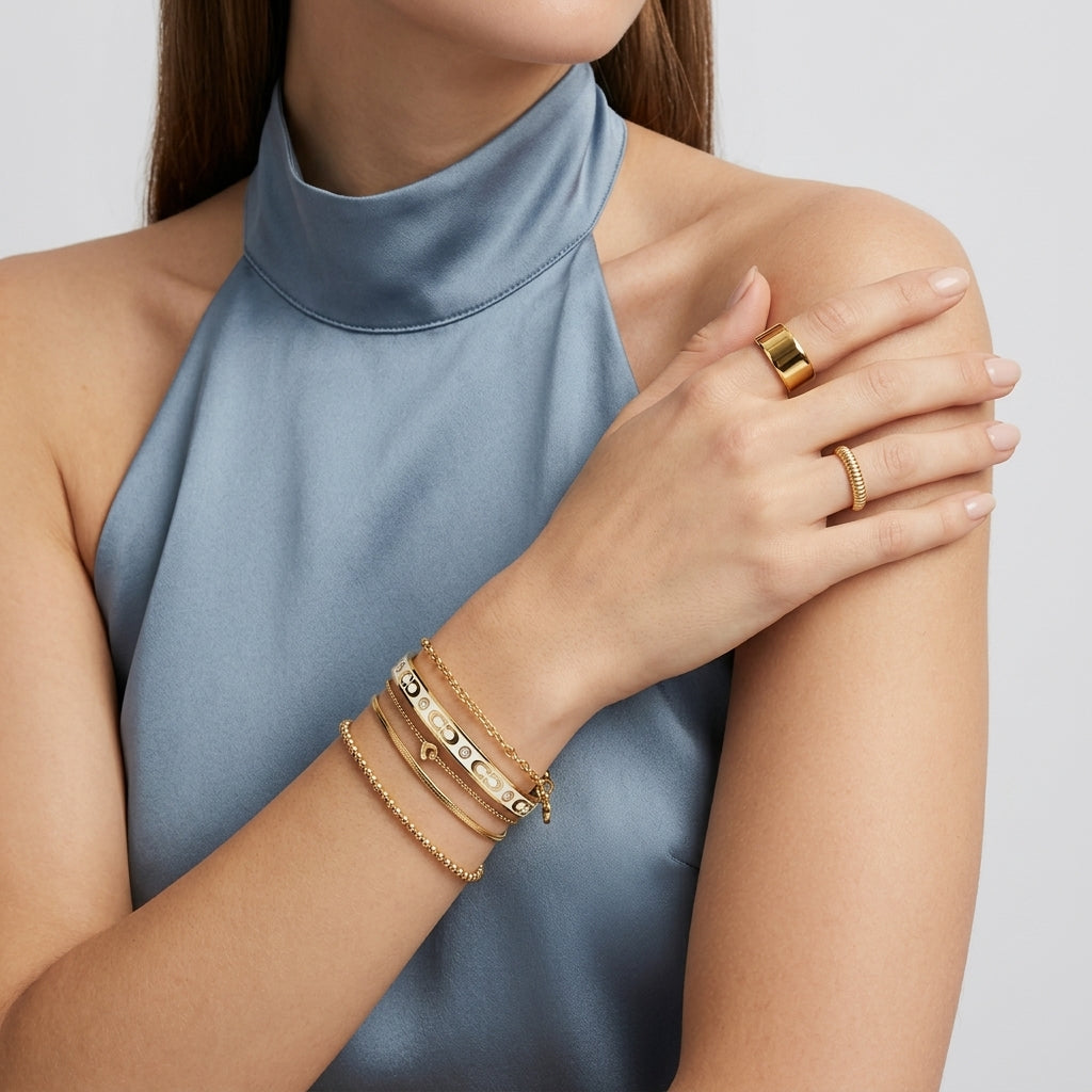 A close-up of a woman wearing delicate gold bracelets and rings on her wrist and hand, paired with a high-neck satin blue top against a light gray background.