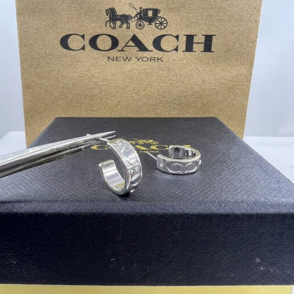 A pair of silver Coach logo hoop earrings displayed on a black presentation box, with a gold Coach shopping bag in the background.