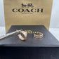 Two Coach New York gold-tone rings with signature horse-and-carriage motifs, displayed on a black box with tweezers in front, and a large Coach New York box behind.