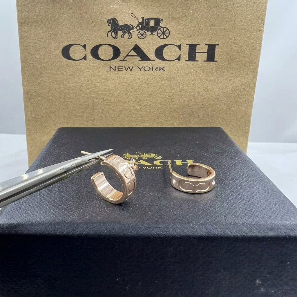 Two Coach New York gold-tone rings with signature horse-and-carriage motifs, displayed on a black box with tweezers in front, and a large Coach New York box behind.