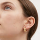 A close-up of a model's ear wearing a small gold huggie hoop earring with a textured, engraved pattern, set against a clean white background.