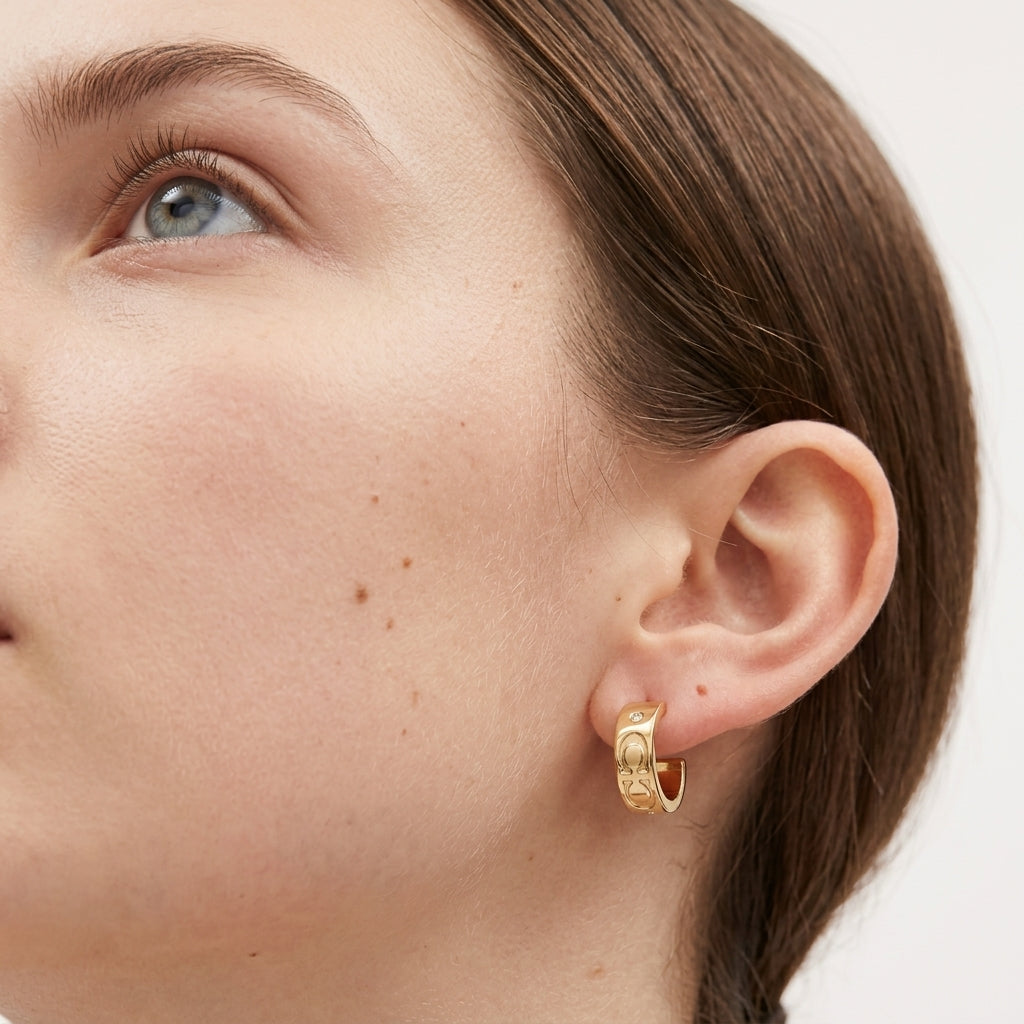 A close-up of a model's ear wearing a small gold huggie hoop earring with a textured, engraved pattern, set against a clean white background.