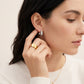 A close-up lifestyle shot of a woman wearing a silver huggie hoop earring and a wide gold band ring, wearing a white knit sweater against a soft beige background.