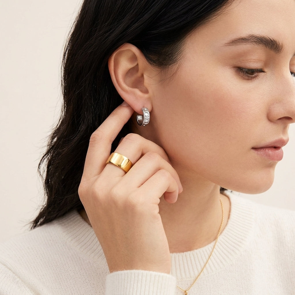 A close-up lifestyle shot of a woman wearing a silver huggie hoop earring and a wide gold band ring, wearing a white knit sweater against a soft beige background.