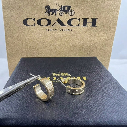 A pair of gold-tone Coach huggie hoop earrings displayed on a black branded box, with tweezers holding one earring open in front of a large Coach New York box.