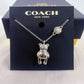 A silver-tone Coach necklace featuring a teddy bear pendant and a pearl accent charm, displayed on a dark navy blue Coach New York branded card insert.