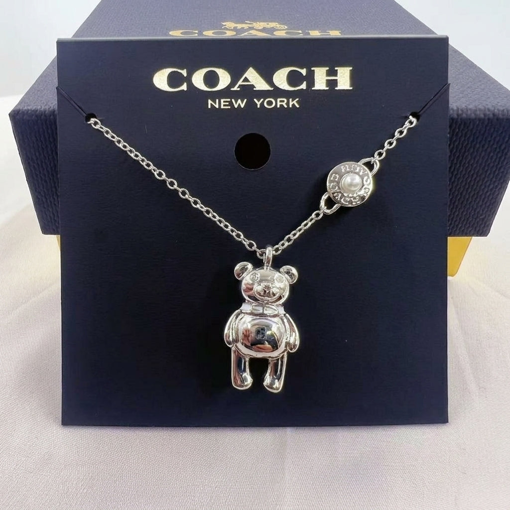 A silver-tone Coach necklace featuring a teddy bear pendant and a pearl accent charm, displayed on a dark navy blue Coach New York branded card insert.
