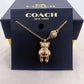 A gold-tone Coach New York necklace with a teddy bear pendant and a pearl accent, displayed on its dark navy blue branded box.
