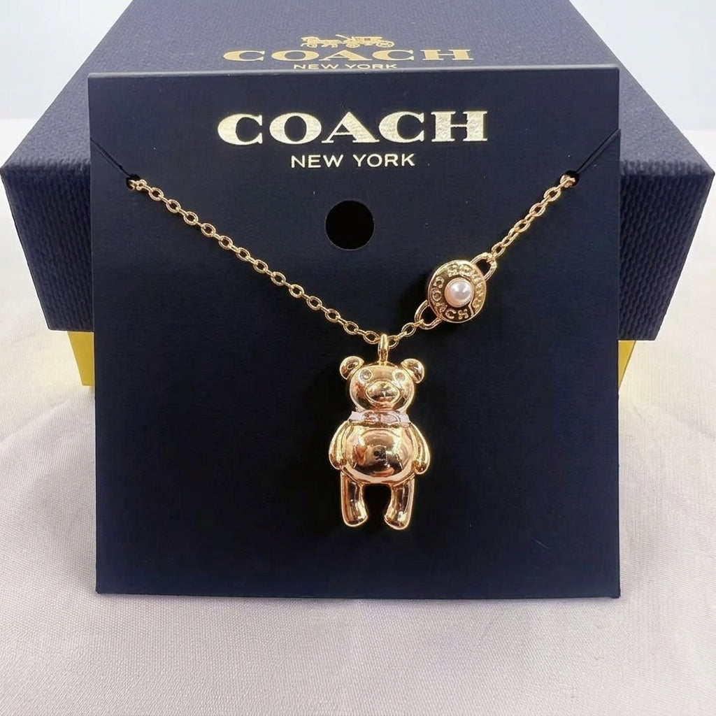 A gold-tone Coach New York necklace with a teddy bear pendant and a pearl accent, displayed on its dark navy blue branded box.