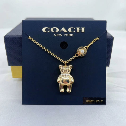 A gold-tone Coach necklace with a teddy bear pendant and a pearl accent, displayed on a dark blue Coach New York branded card against a white background.