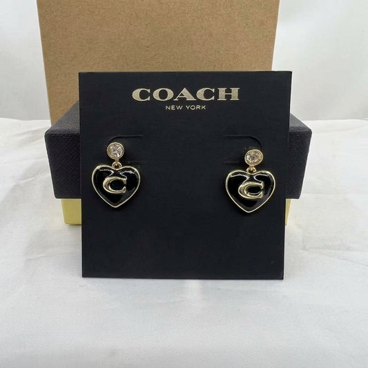 A pair of Coach New York heart-shaped drop earrings with gold-tone metal, a clear crystal stud, and a raised 'C' logo, displayed on a black branded card insert.