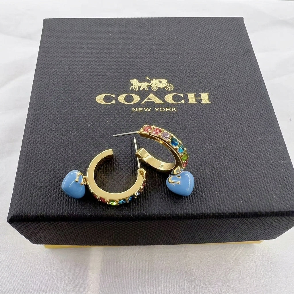 Gold hoop earrings with blue heart-shaped dangles and multicolor rhinestone trim, displayed on a black Coach New York box.