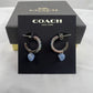 A pair of Coach New York hoop earrings displayed on a black branded card, with the textured black Coach box behind it. The earrings have silver-toned hoops with multicolor rhinestones and light blue heart-shaped charms.