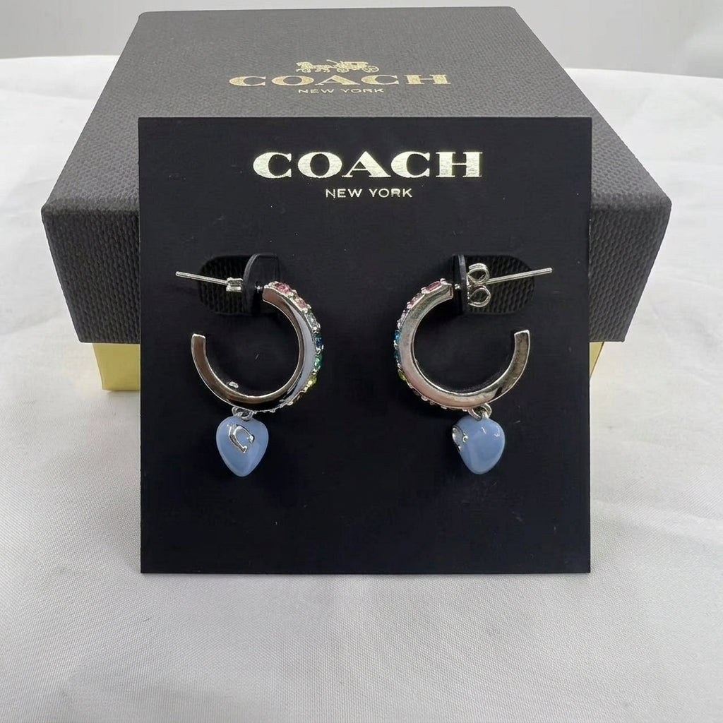 A pair of Coach New York hoop earrings displayed on a black branded card, with the textured black Coach box behind it. The earrings have silver-toned hoops with multicolor rhinestones and light blue heart-shaped charms.