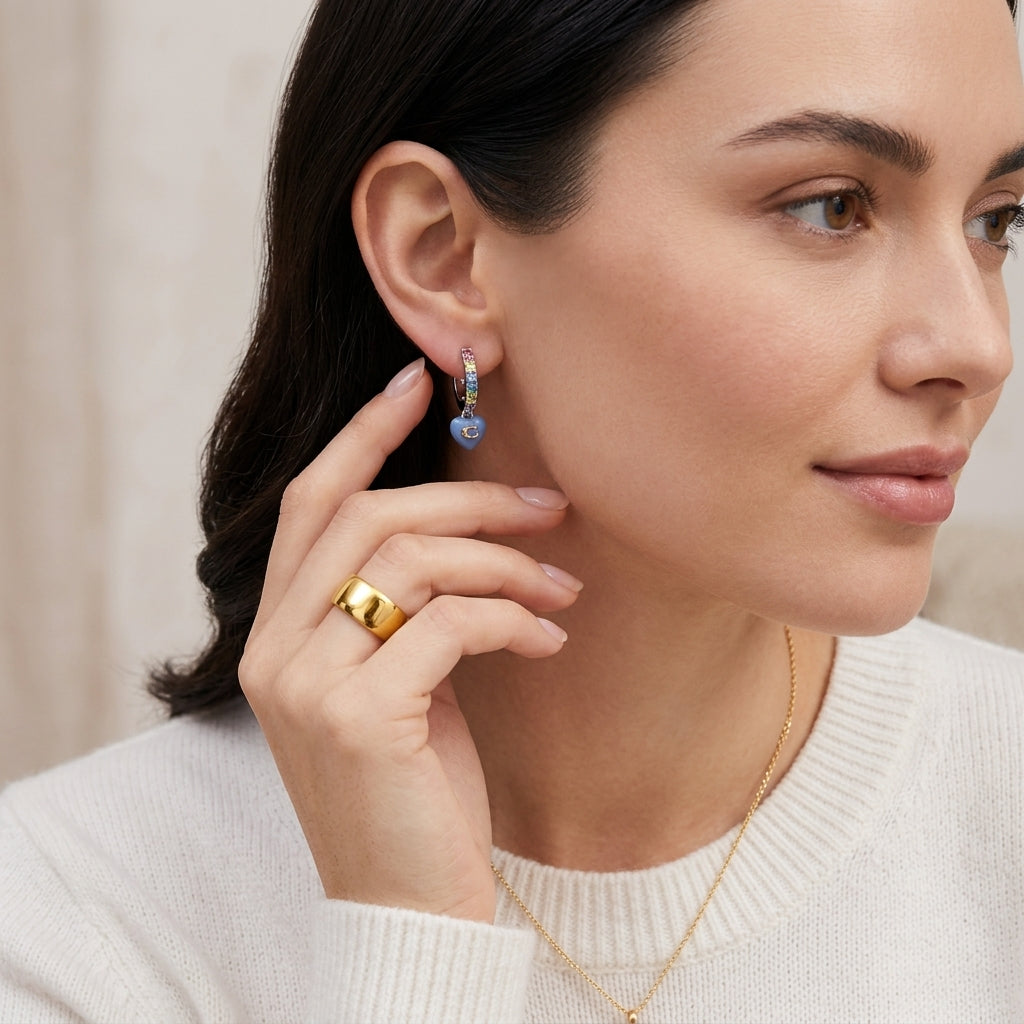 A close-up lifestyle shot of a woman wearing a small gold hoop earring with a blue heart-shaped stone, along with a gold ring and necklace.