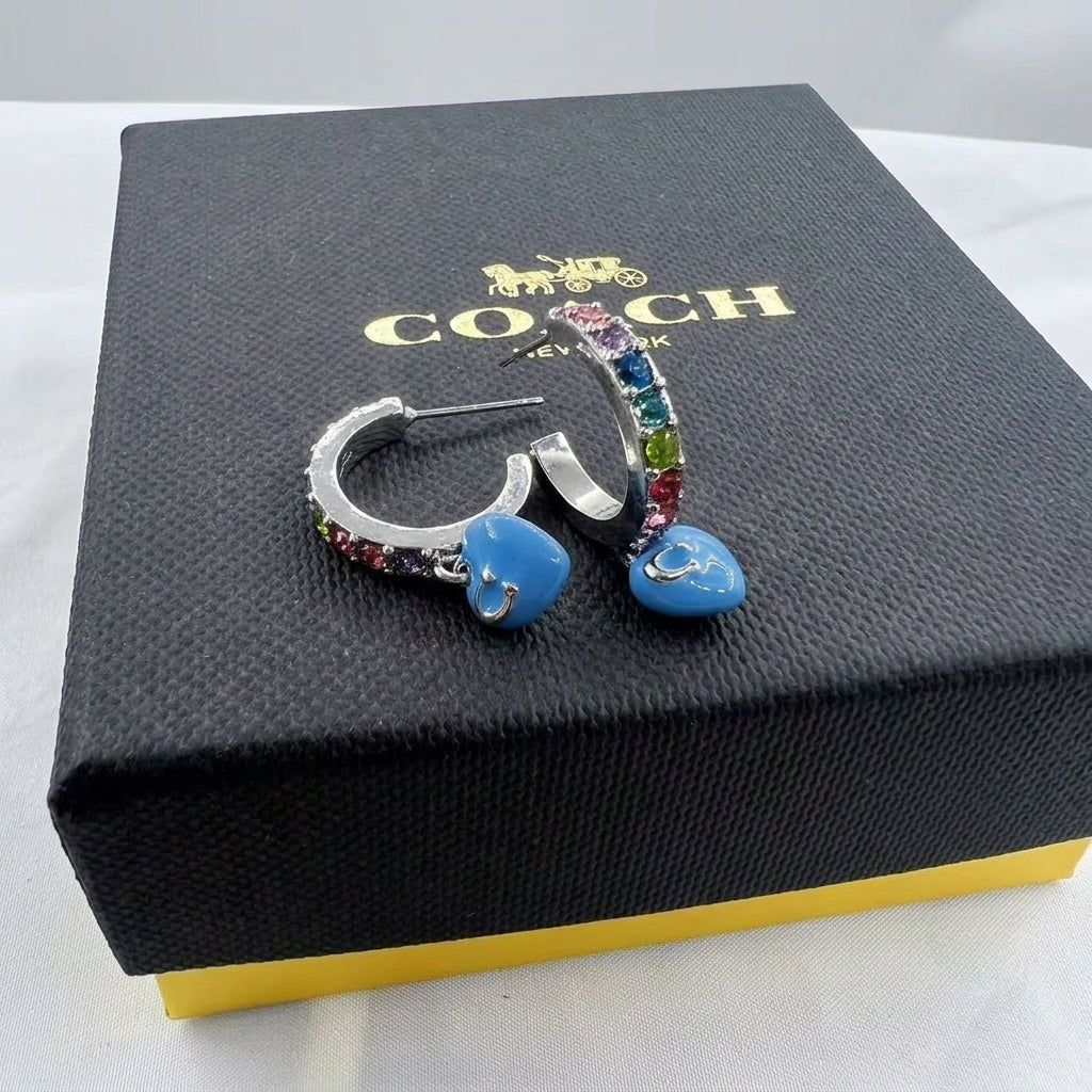 A pair of Coach New York hoop earrings with colorful rhinestone accents and blue heart charms, displayed on top of a black Coach New York box.