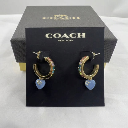A pair of Coach New York gold hoop earrings with colorful rhinestones and a dangling light blue heart charm, displayed on a black branded card in front of a dark gray Coach box.