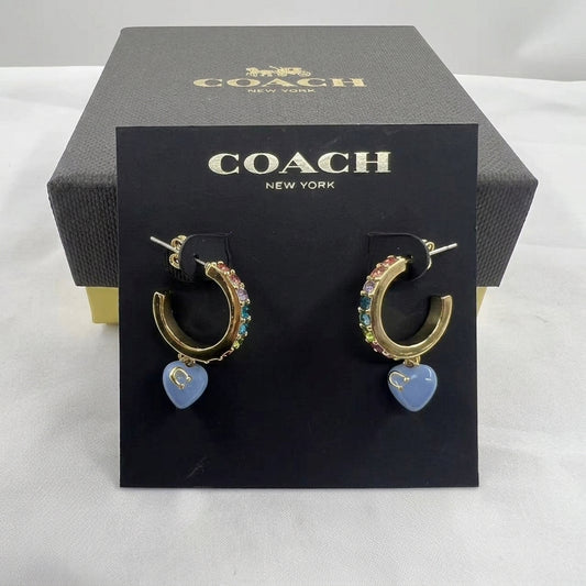 A pair of Coach New York gold hoop earrings with colorful rhinestones and a dangling light blue heart charm, displayed on a black branded card in front of a dark gray Coach box.