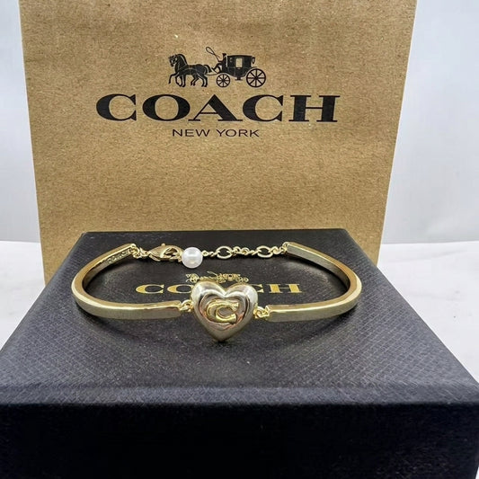 A gold-tone Coach bracelet with a heart-shaped logo charm and a small pearl accent, displayed on a black Coach box with a brown Coach New York shopping bag behind it.