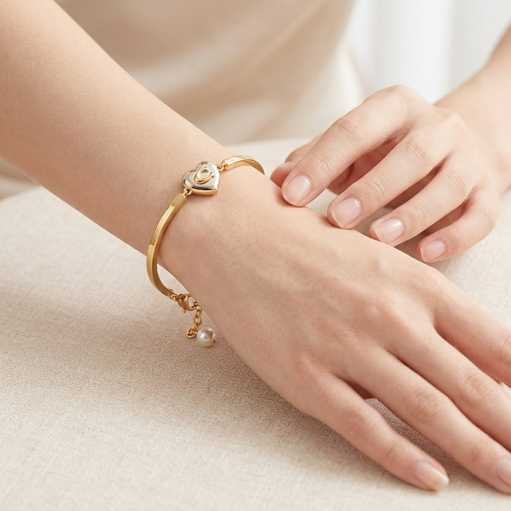 A gold bangle bracelet with a heart-shaped charm and a small dangling pearl, shown worn on a person's wrist with hands resting on a light beige fabric surface.