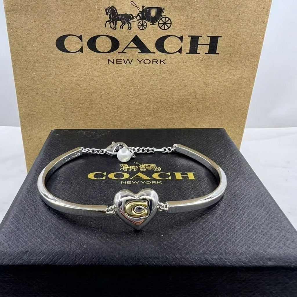 A silver-tone Coach bracelet with a heart-shaped charm featuring a gold 'C' logo, displayed on a black Coach box with the brand's signature box behind it.