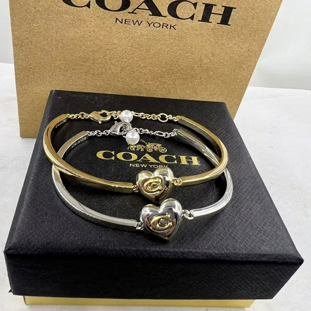 A Coach New York bracelet set displayed on a black presentation box with a kraft Coach box behind it. The set includes a gold bangle with a rose and pearl charm, a silver bangle with a heart charm, and a gold-toned chain link bracelet.