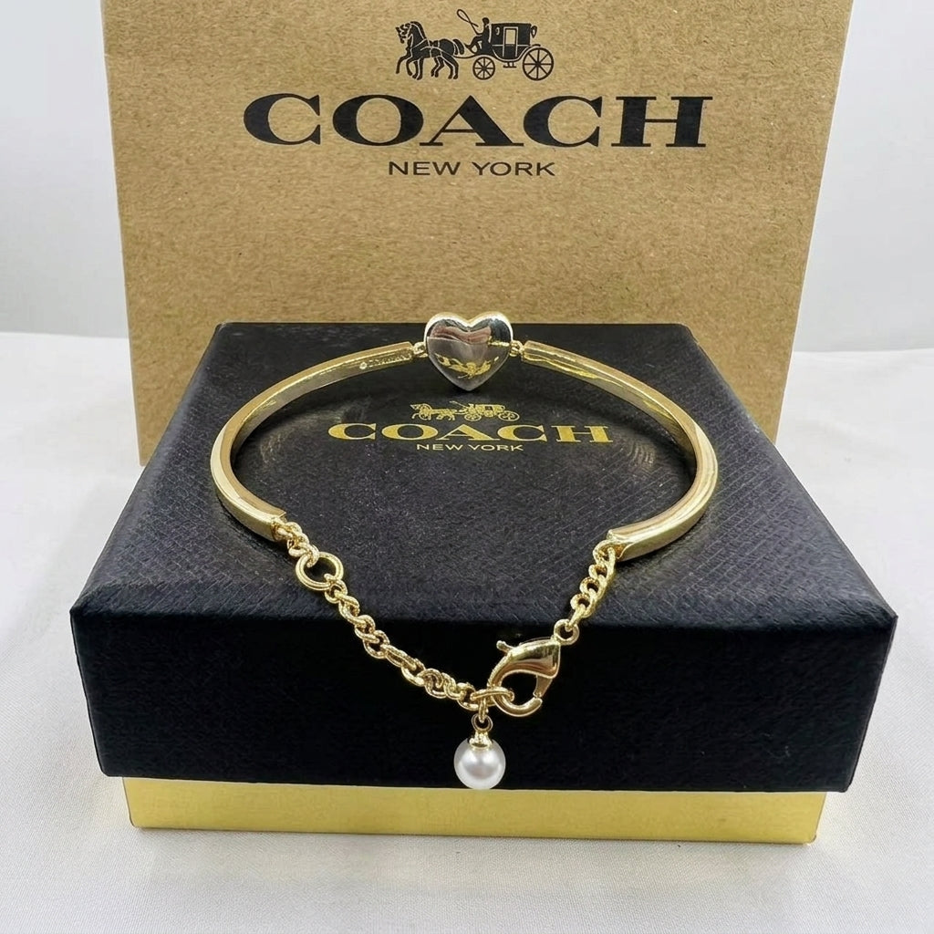 A gold-tone Coach bangle bracelet with a heart charm and a dangling pearl, displayed on a black Coach box with a tan Coach New York box behind it.