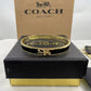 A gold-tone Coach hinged bangle bracelet with a horse-and-carriage charm, displayed on a black presentation box with a brown Coach box behind it.