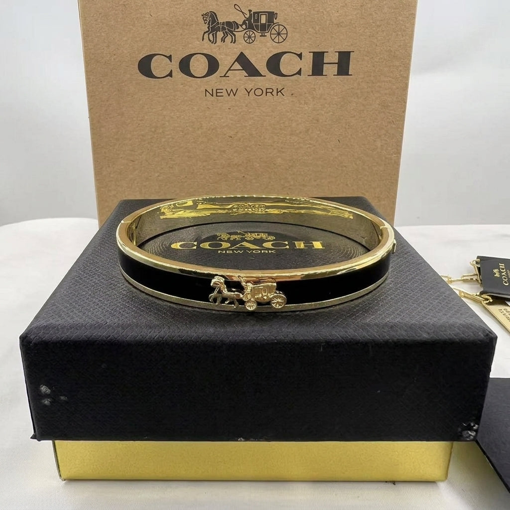 A gold-tone Coach hinged bangle bracelet with a horse-and-carriage charm, displayed on a black presentation box with a brown Coach box behind it.