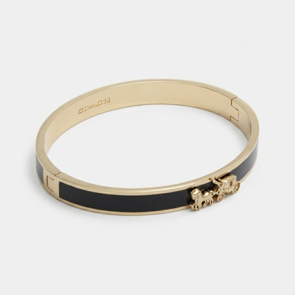 A gold-tone Coach bangle bracelet with a black enamel inlay and a small horse-and-carriage charm, shown on a light gray background.