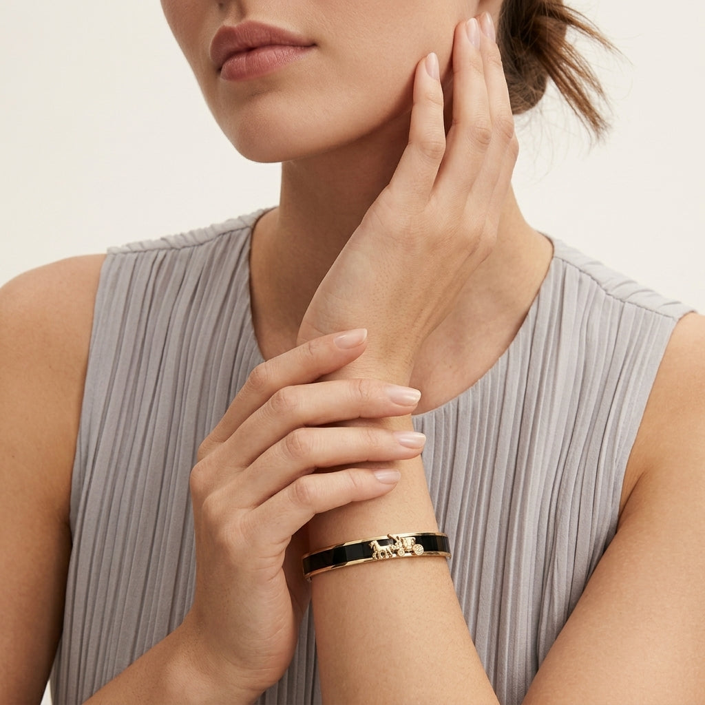 A close-up of a woman wearing a slim black bangle bracelet with gold-toned lettering that reads 'LOVE YOU'. She is wearing a light gray pleated top against a clean, off-white background.