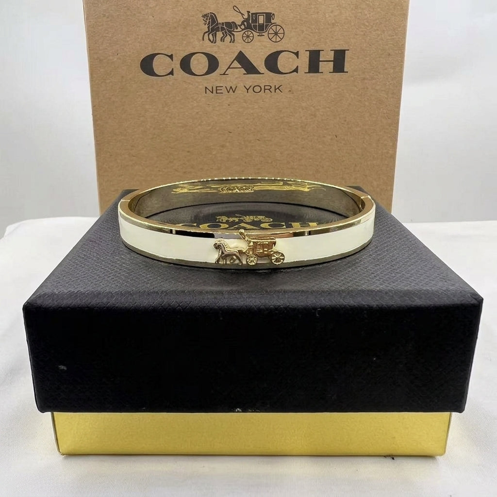 A Coach New York gold-tone bangle bracelet with an off-white enamel inlay and a horse carriage logo, displayed on a black presentation box with its brown cardboard box behind it.