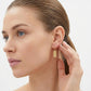 A close-up side profile of a woman with her hair pulled back, wearing a small gold dangle earring with a textured rectangular charm, against a clean white background.