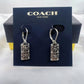 A pair of silver-tone Coach drop earrings with rectangular interlocking logo charms, displayed on a dark navy blue Coach New York branded card, with the Coach box visible in the background.