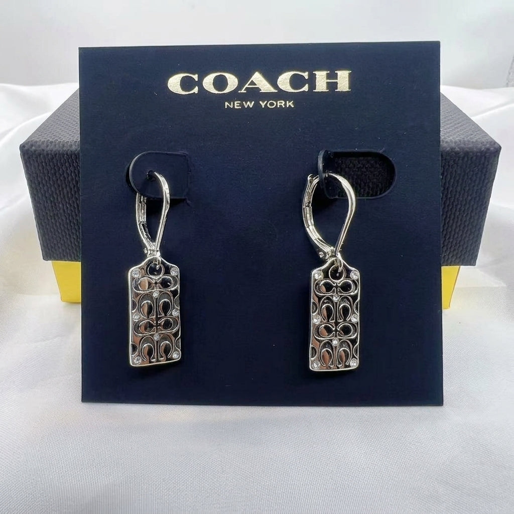 A pair of silver-tone Coach drop earrings with rectangular interlocking logo charms, displayed on a dark navy blue Coach New York branded card, with the Coach box visible in the background.