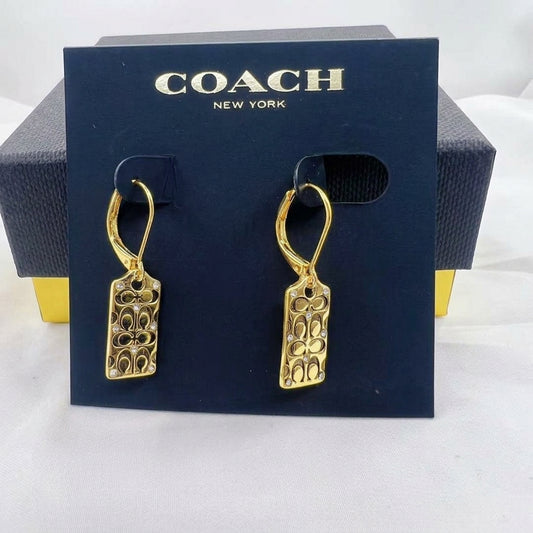 A pair of gold-tone Coach drop earrings with a signature 'C' pattern, displayed on a dark blue Coach New York branded card, with the original dark blue box partially visible behind it.