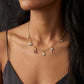 A close-up of a model wearing a delicate gold chain necklace with small charms including a flower, key, and animal motifs, styled with a dark camisole.