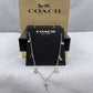 A silver-tone Coach necklace with charms including a carriage, star, bird, and key, displayed on a black Coach card with the Coach New York box behind it.