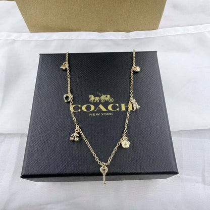 A gold-tone Coach necklace with multiple charm motifs, including a key at the center, displayed on top of a black Coach New York box.