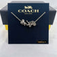 A silver-tone Coach New York necklace with a horse-drawn carriage pendant, displayed on its dark navy blue branded box.
